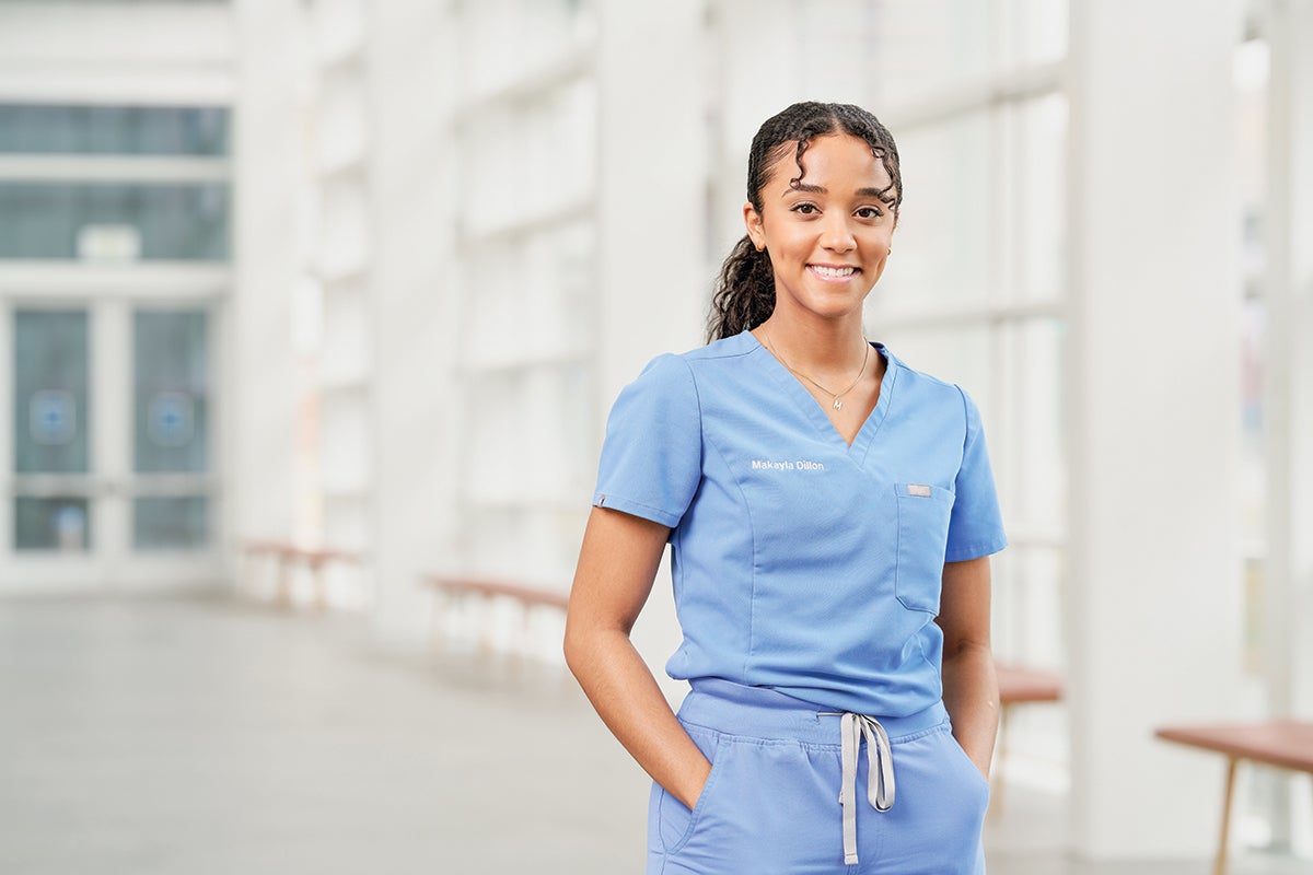 An image of a dental student standing smiling in blue scrubs and in a long window-filled corridor.