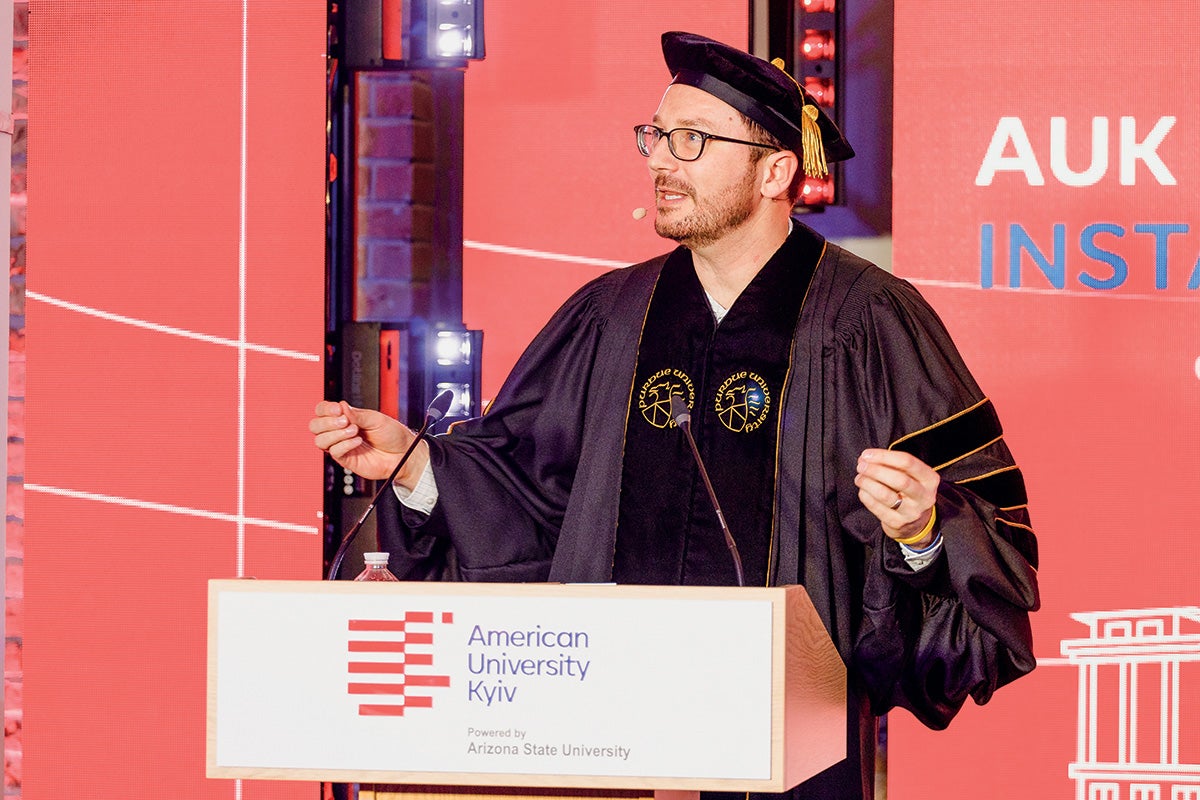 A photo of Roman Sheremeta speaking at a podium and welcoming students at the start of the inaugural year of American University Kyiv. He is wearing a cap and gown.