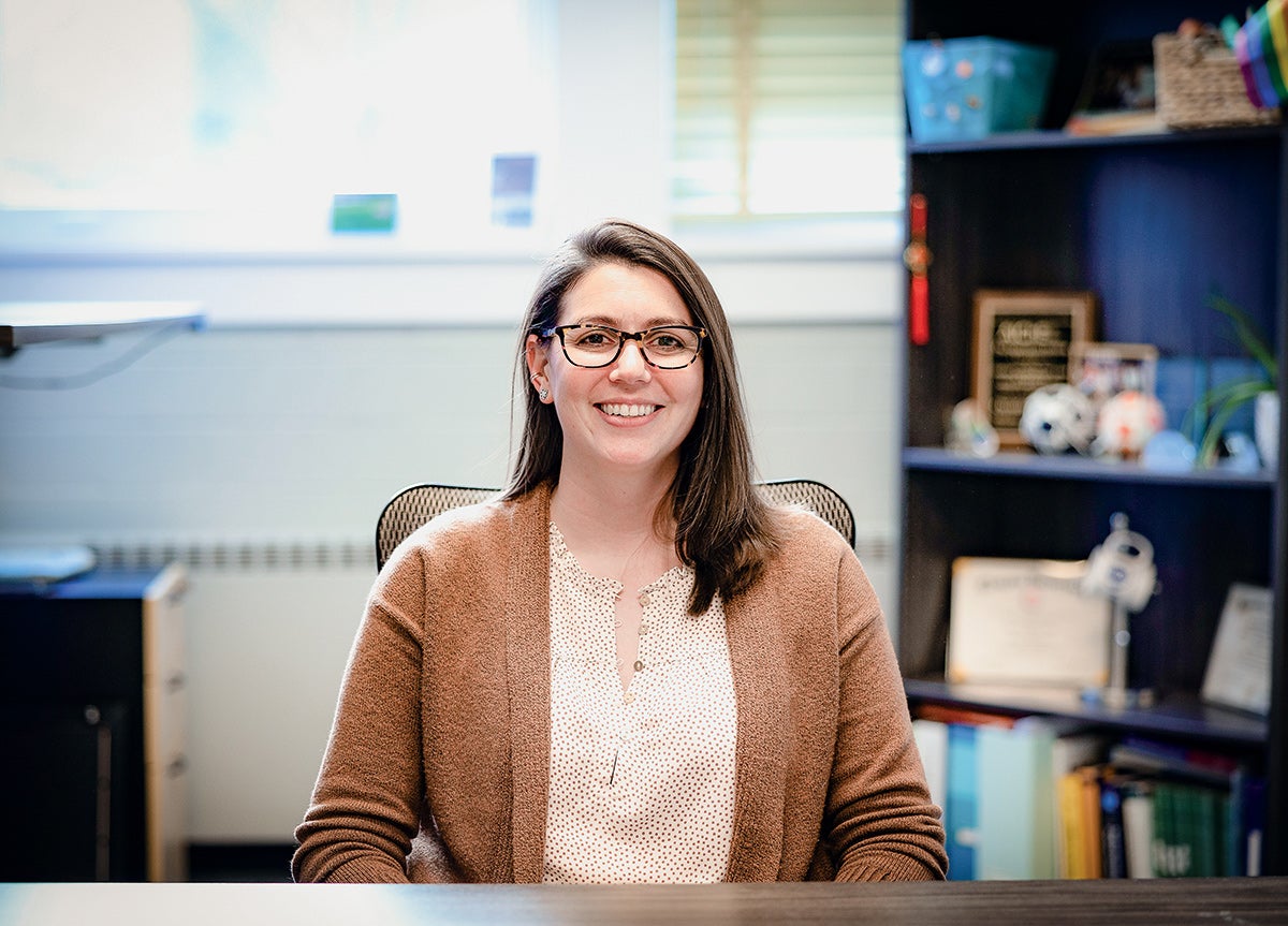 Professor Christine Duval sitting at her desk in her campus office with a bookshelf to her side.