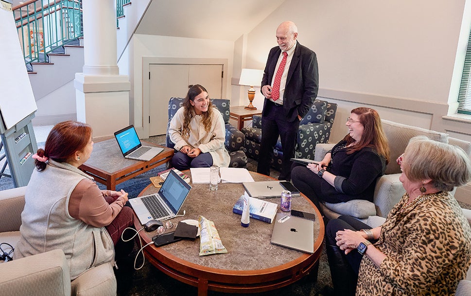 Professor Frank Barrett stands and talks with several students sitting in living room-like chairs around a table.