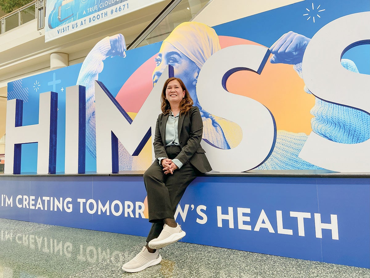 Mary Joy Garcia-Dia casually sits in front of a large sign for the Healthcare Information and Management Systems Society conference, where she spoke.