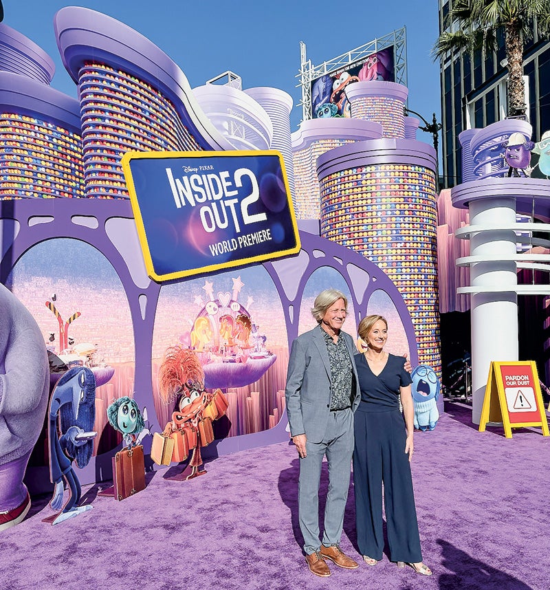 Image of two people standing in front of a sign for the animated movie, 'Inside Out 2' sign, and on the purple carpet for the movie's opening.