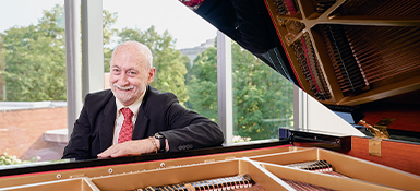 Professor Frank Barrett, who also is a pianist, sitting at a piano bench, framed by the piano and the raised lid.