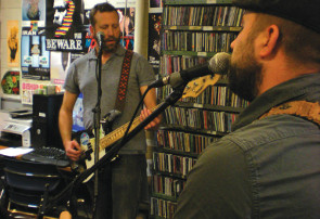Two bearded men with guitars behind mics and in front of a wall of CDs.