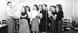 B&W--a male student directing several singing female students in front of mics.