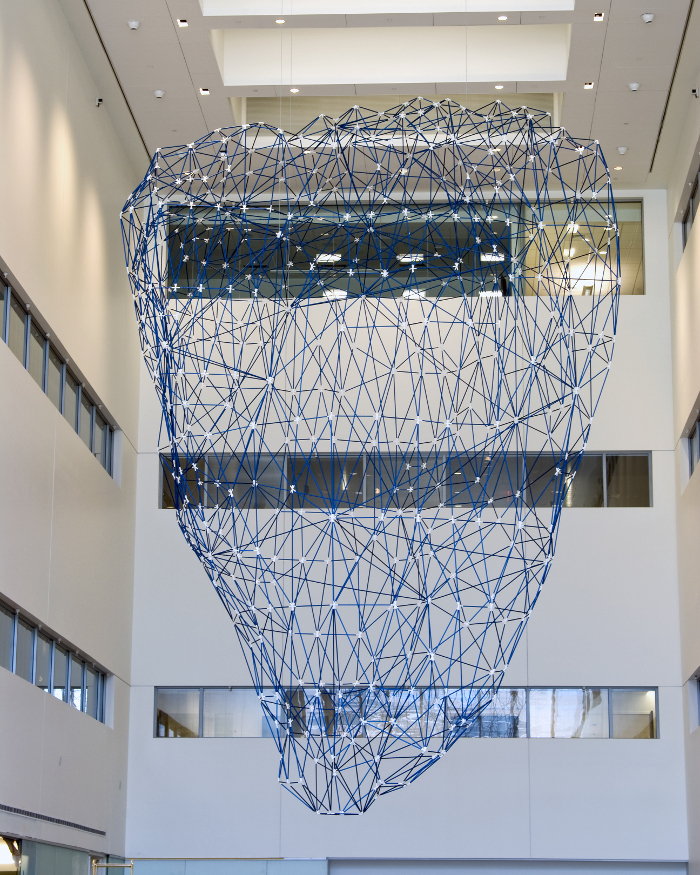 A piece of abstract art that is being hung from the ceiling. The sculpture is made out of blue aluminum pieces and is modelled after an iceberg