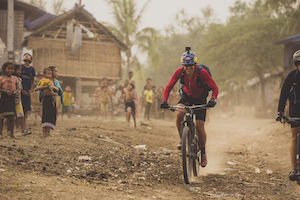 Photo of a woman, Rebecca Rusch, riding a bike through a village on the Ho Chi Minh trail, with many children watching