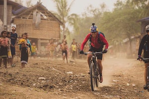 Photo of a woman, Rebecca Rusch, riding a bike through a village on the Ho Chi Minh trail, with many children watching