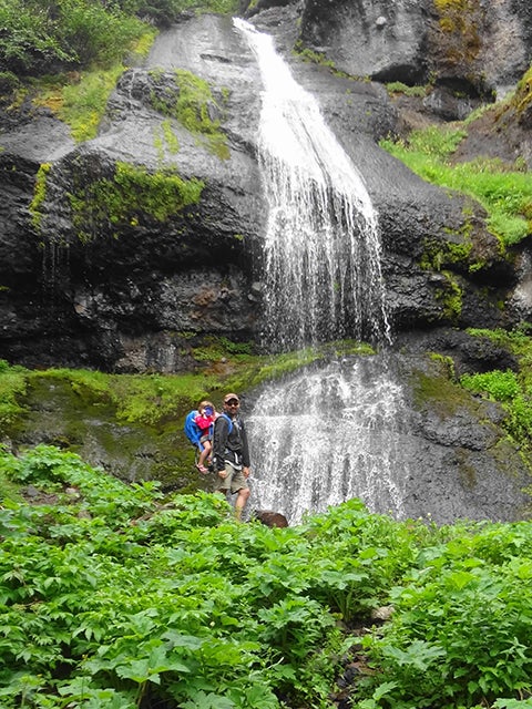 Jonathan Adler standing in front of a waterfall with his daughter harnessed to his back