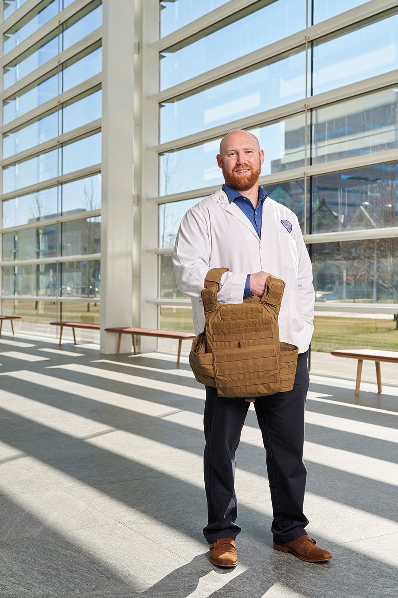 Medical student Luke Flint in a white coat holding a bullet-proof vest he wore in the U.S. air force