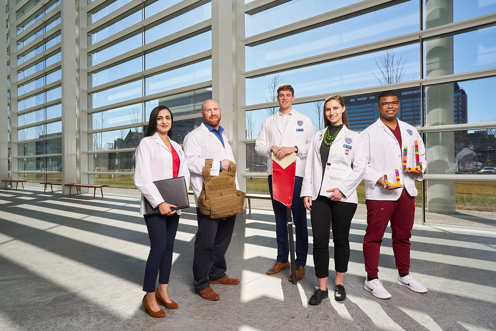 Five medical students in white coats and holding objects from their past work in front of a large set of windows.