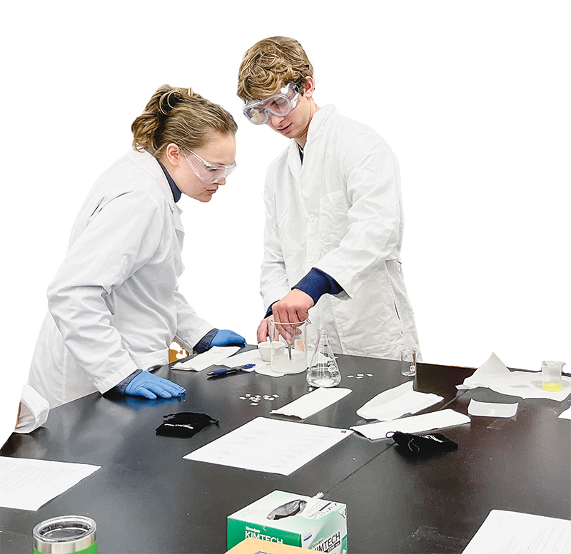 Two students working on a chemical experiment, wearing lab coats and goggles