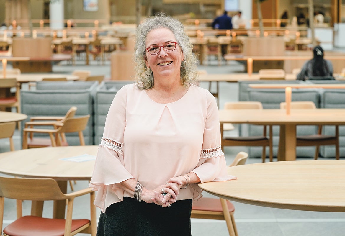 Photo of Susan Painter standing, smiling in a lobby with tables and chairs behind her.