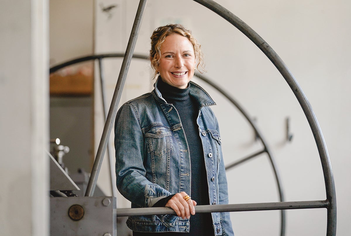 AA photo of art historian Erin Benay standing next to the large wheel of a press used by printmakers.
