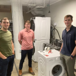 Three young men stand around a washing machine.