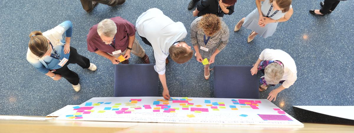 An overhead photo of seven individuals wearing conference badges standing before a white board covered in colorful post-it notes during a brainstorming session.