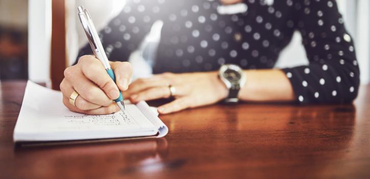 Shot of a unrecognizable woman writing in a book with a pen on a dinner table at home