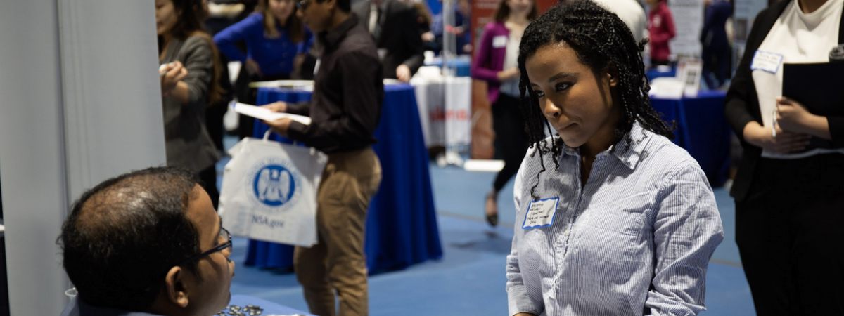 A student of color speaks with an employer sitting at a table at a career fair