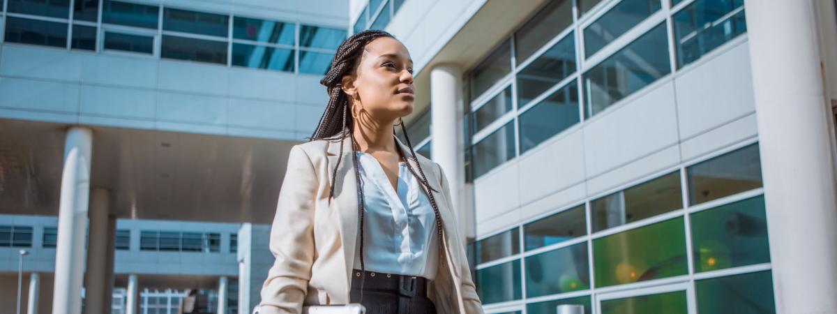 A woman of color in business attire stands regally outside a modern hospital complex