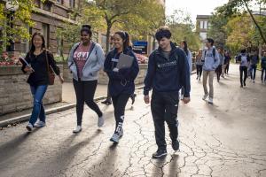 Four students walk on campus with the backpacks and laptops on a fall day.