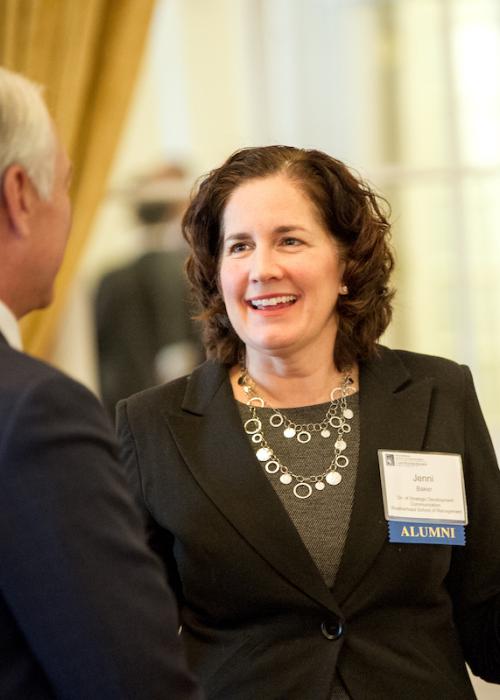 Alumna Jenni Baker stands wearing a name tag as she smiles and gestures in conversation at the Bowers Luncheon event