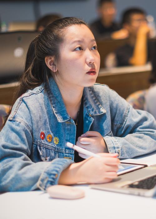 A student of Asian descent sitting in a lecture hall looks to professor and takes notes