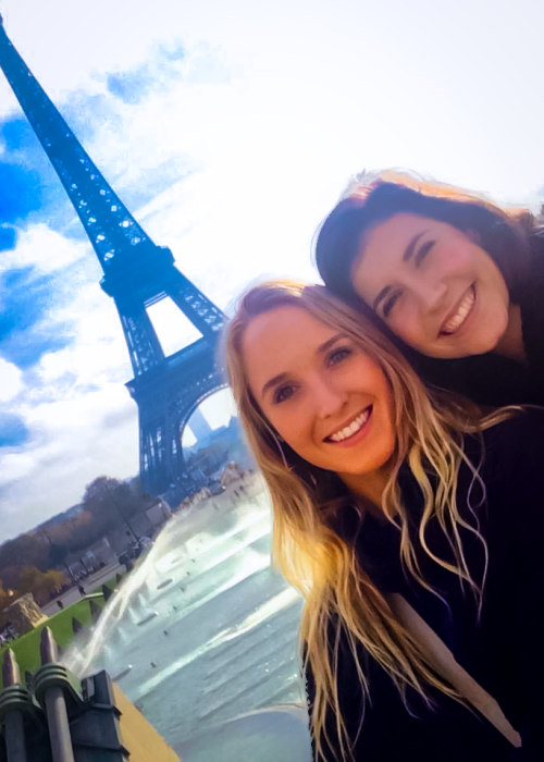 Two femme-presenting students pose together smiling with the Eiffel Tower and blue sky in the background