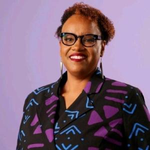 Headshot of Hermione Malone, who is wearing black framed glasses and a bold, patterned blazer with a mix of black, purple, and blue geometric designs.