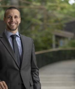 Jude Klak smiles outside surrounded by greenery in a suit and tie.