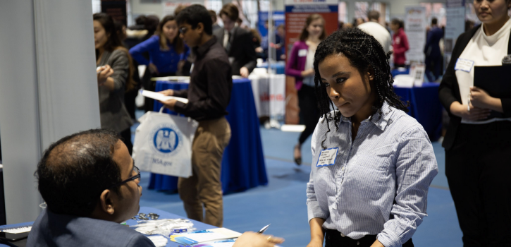A student of color speaks with an employer sitting at a table at a career fair