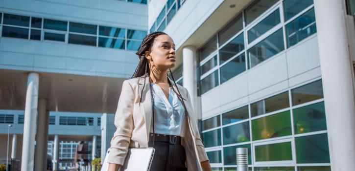 A woman of color in business attire stands regally outside a modern hospital complex