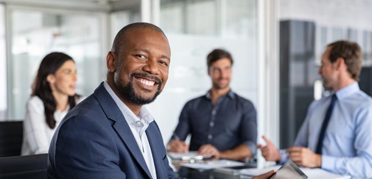 A stock photo of a business professional smiling at the camera while his coworkers sit in a meeting
