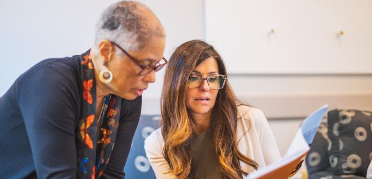One person stands over a seated person as they both review paperwork together.