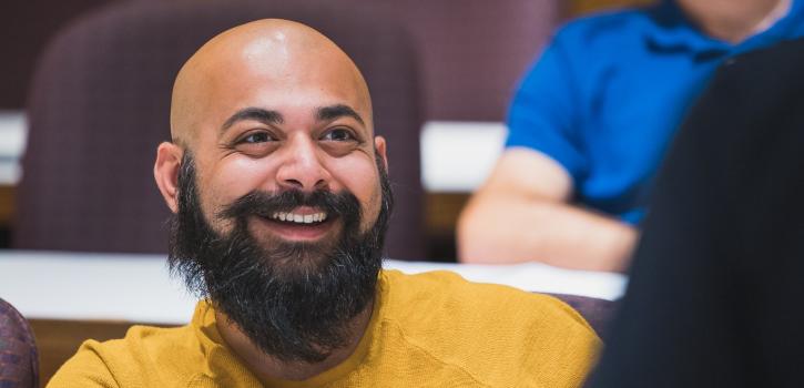 A student with a beard and a yellow shirt smiling during a lecture.