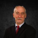 This image features Frank Barrett, who is wearing a black suit, a light blue dress shirt, and a red tie with a subtle pattern.