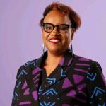 Headshot of Hermione Malone, who is wearing black framed glasses and a bold, patterned blazer with a mix of black, purple, and blue geometric designs.