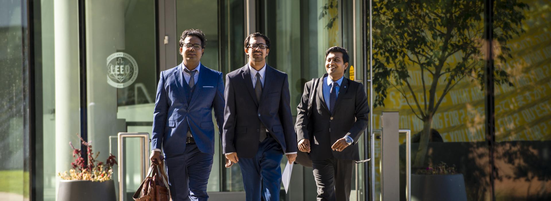 Three students dressed in business atire walk out of a building with briefcases. 