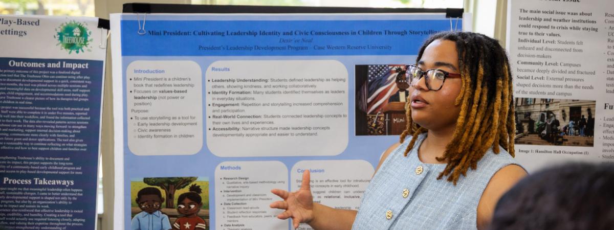 A young woman with glasses gestures toward a blue research poster, presenting at an academic event at Case Western Reserve University.