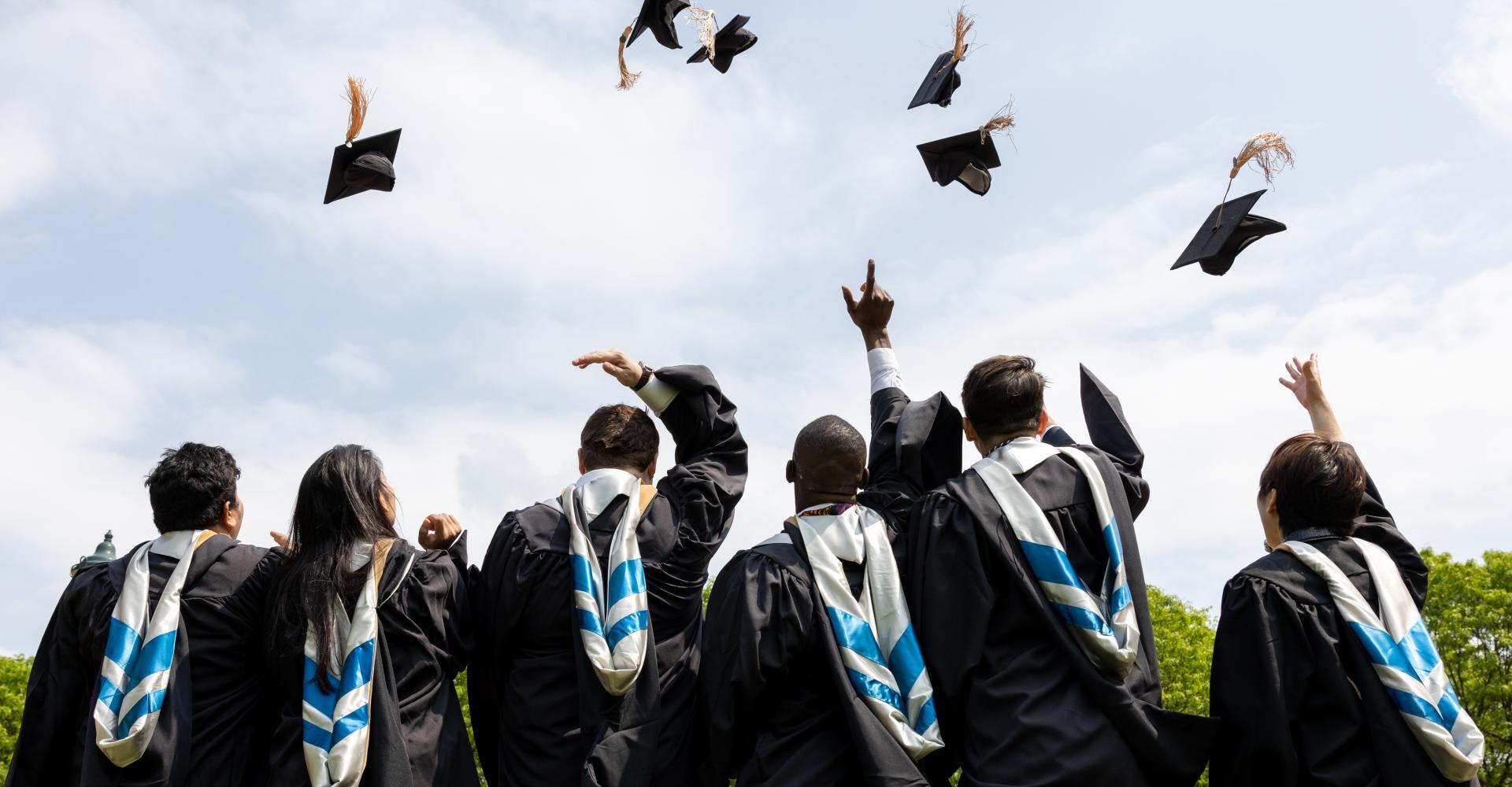 Six college graduates with their backs to the camera while throwing their graduation caps in the air