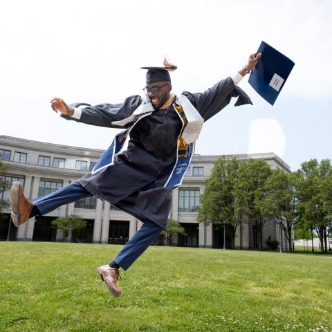 Student in graduation attire in an outdoor setting kicking his heels while jumping in the air.
