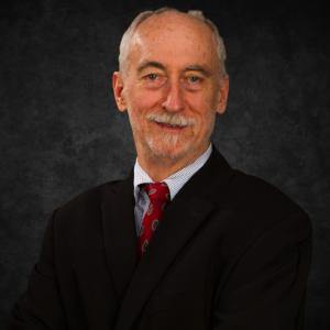 Frank Barrett smiles in a suit against a dark background.