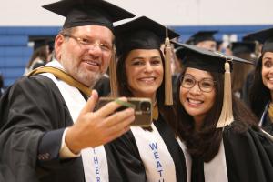 Three students in graduation attire in an outdoor taking a selfie photo