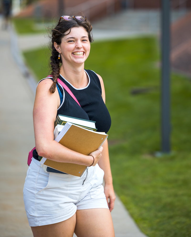 Student standing outdoors smiling with hair in a braid while holding a stack of books by one arm