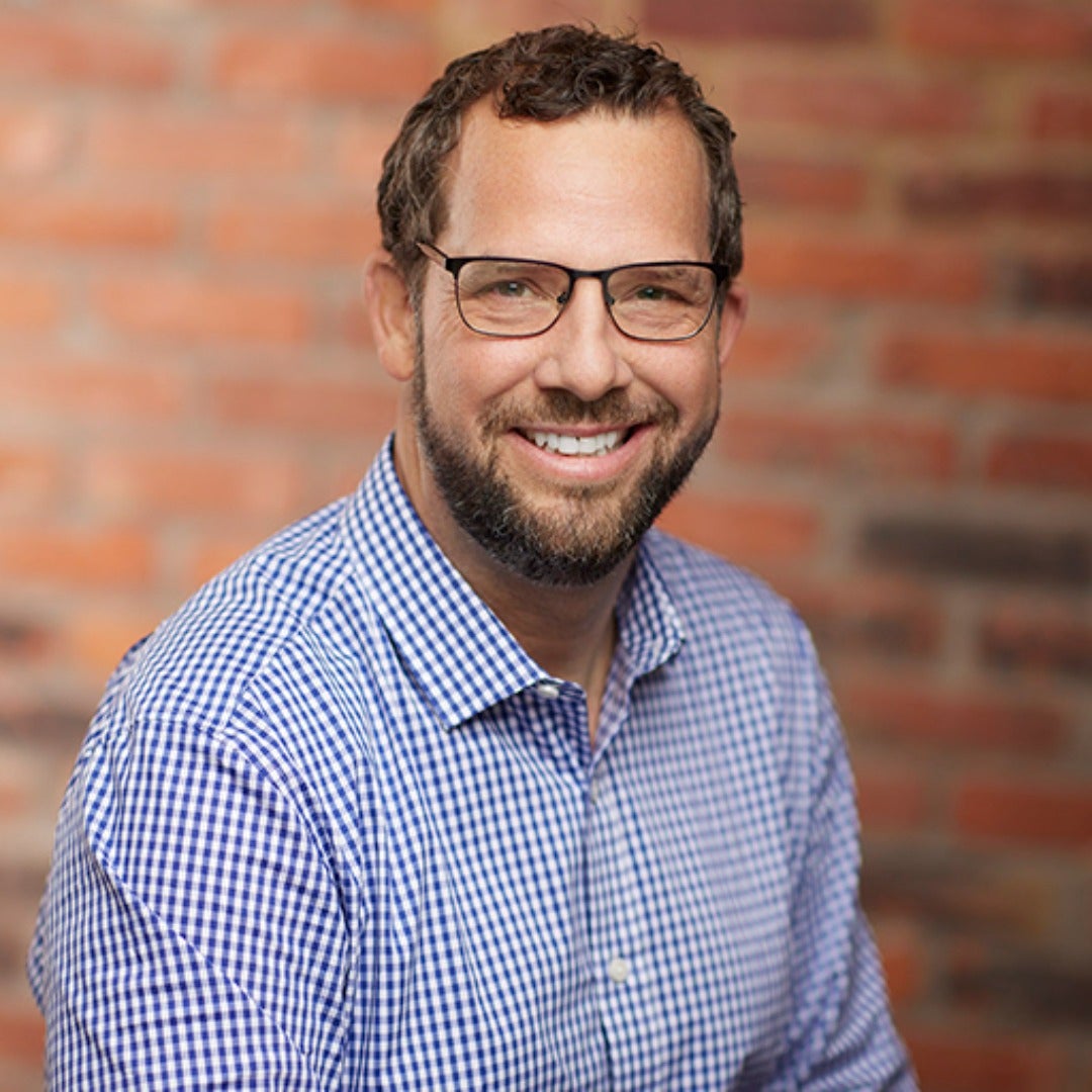 Scot Lowry smiles in a button up shirt against a red brick background.