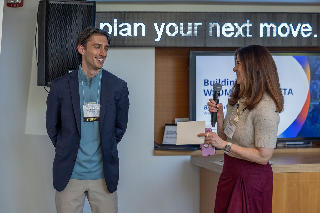 A smiling student stands next to a woman speaking into a microphone and holding a small note card during an event at the Weatherhead School of Management.