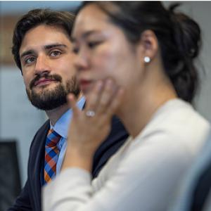 person sitting at table with suit on next to another person looking at a computer