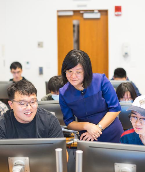 A faculty instructor of color looks at the computer screens of two students of color during class