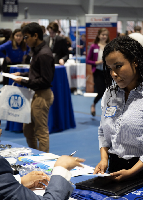 A student of color speaks with an employer sitting at a table at a career fair