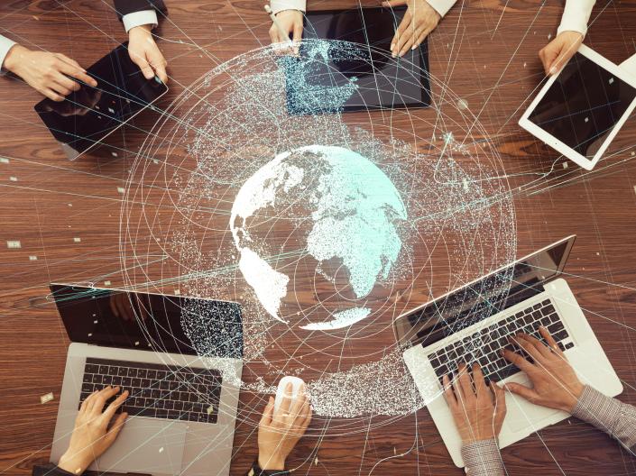 Birds-eye-view of 5 people's hands using laptops and tablets; Centered is a semi-translucent graphic of the globe 