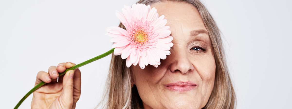 Close up of a woman's face. She holds a pink flower over one eye